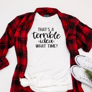 K&ouml;nnte beinhalten: Wei&szlig;es T-Shirt mit schwarzem Text, der "That's a terrible idea - what time?" lautet. Das T-Shirt liegt flach auf einer wei&szlig;en Oberfl&auml;che, dar&uuml;ber liegt ein rot-schwarzes Karohemd.