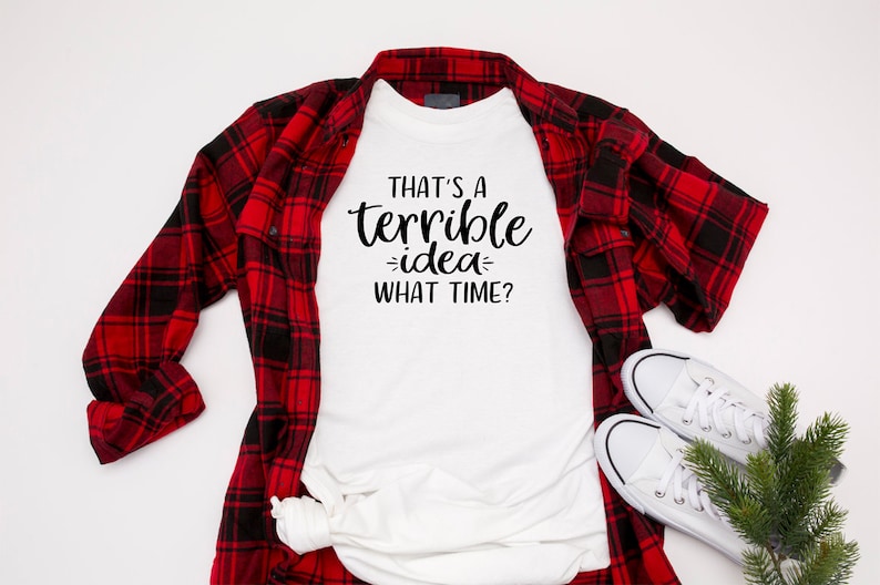 K&ouml;nnte beinhalten: Wei&szlig;es T-Shirt mit schwarzem Text, der "That's a terrible idea - what time?" lautet. Das T-Shirt liegt flach auf einer wei&szlig;en Oberfl&auml;che, dar&uuml;ber liegt ein rot-schwarzes Karohemd.
