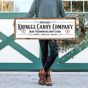 Pu&ograve; includere: Un cartello rettangolare in legno con sfondo bianco e cornice marrone scuro. Il testo recita: OPEN DAILY KRINGLE CANDY COMPANY OLDE FASHIONED CANDY CANES AUTHENTIC HAND ROLLED MADE DAILY. Sono incluse due piccole illustrazioni di bastoncini di zucchero.
