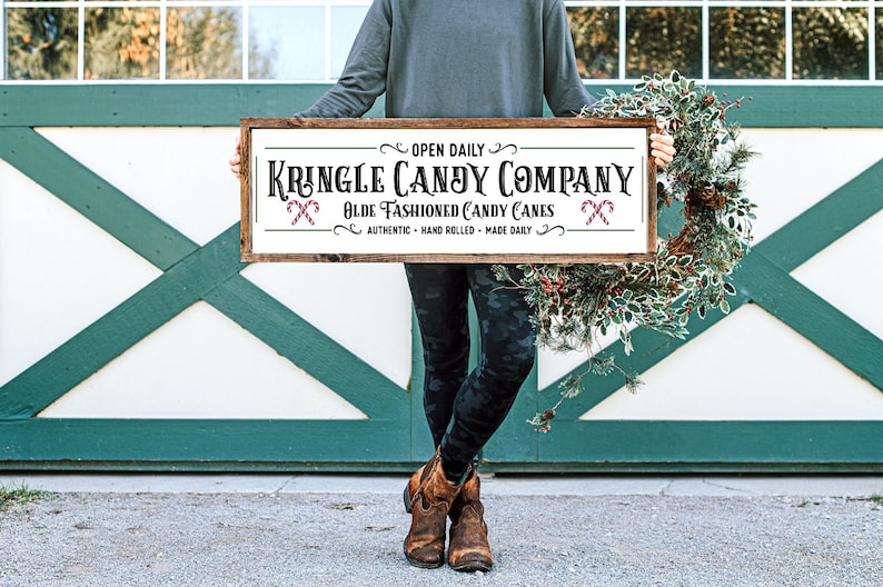 Pu&ograve; includere: Un cartello rettangolare in legno con sfondo bianco e cornice marrone scuro. Il testo recita: OPEN DAILY KRINGLE CANDY COMPANY OLDE FASHIONED CANDY CANES AUTHENTIC HAND ROLLED MADE DAILY. Sono incluse due piccole illustrazioni di bastoncini di zucchero.
