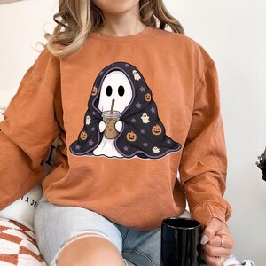 Spooky season digital PNG with a cozy ghost, iced drink, black Halloween blanket, orange pumpkins, cream ghosts, and purple star details.
