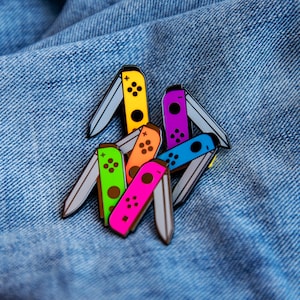 Puede incluir: Un conjunto de siete pines de esmalte de colores con forma de mandos de Nintendo Switch. Los pines están dispuestos en una pila sobre un fondo de mezclilla azul. Los colores de los pines son amarillo, naranja, verde, rosa, azul, morado y gris.