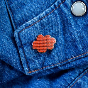May include: A red brick cloud enamel pin on a blue denim background.