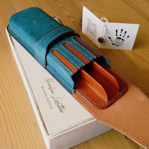 May include: A teal blue leather pen case with brown leather interior and stitching. The case has three slots for pens and a flap that closes with a snap. The case is handcrafted and has the words "Genuine Leather Handcrafted" embossed on the front.