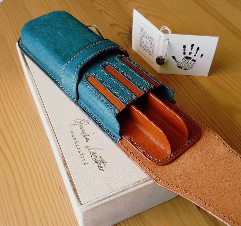 May include: A teal blue leather pen case with brown leather interior and stitching. The case has three slots for pens and a flap that closes with a snap. The case is handcrafted and has the words "Genuine Leather Handcrafted" embossed on the front.