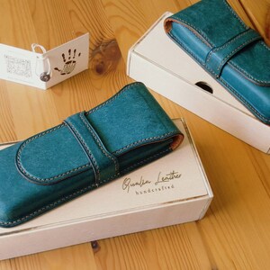 May include: Two teal blue leather pen cases with a brown leather strap and stitching. The cases are on top of white boxes with the text "Qualia Leather handcrafted" printed on the box.