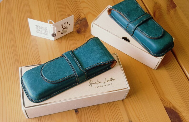 May include: Two teal blue leather pen cases with a brown leather strap and stitching. The cases are on top of white boxes with the text "Qualia Leather handcrafted" printed on the box.