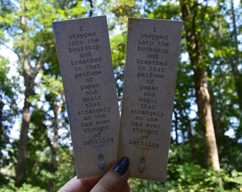 May include: Two brown paper bookmarks with a blue and gold design. The bookmarks feature a quote about the smell of books: "I stepped into the bookshop and breathed in that perfume of paper and magic that strangely no one had ever thought of bottling."