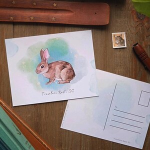 May include: A watercolor postcard featuring a brown rabbit with a white belly, set against a blue and green watercolor background. The postcard reads "Travelers Rest, SC".