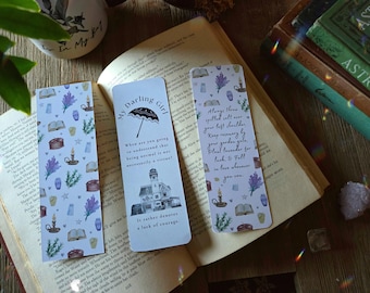 May include: Three decorative bookmarks with whimsical designs and text, resting on an open book. One bookmark reads "My Darling Girl" with an umbrella illustration. The other two feature botanical and celestial patterns with quotes.