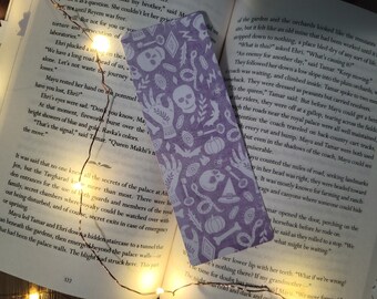 May include: A lavender bookmark with a repeating white pattern of skulls, hands, keys, and other mystical symbols. The bookmark is placed in an open book with text visible.