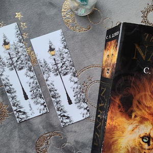 Könnte beinhalten: Zwei schwarz-weiße Lesezeichen mit einer goldenen Laterne und schneebedeckten Bäumen. Die Lesezeichen befinden sich auf einem grauen Hintergrund mit goldenen Sternen. Der Text auf dem Lesezeichen lautet "The Narnian".