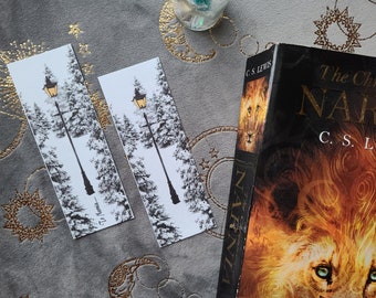 May include: Two black and white bookmarks featuring a gold lamppost and snowy trees. The bookmarks are on a grey background with gold stars. The text on the bookmark reads "The Narnian".