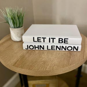 May include: Two white books stacked on a wooden table. The top book reads "LET IT BE" in black letters, and the bottom book reads "JOHN LENNON". A small potted plant with green grass sits to the left.