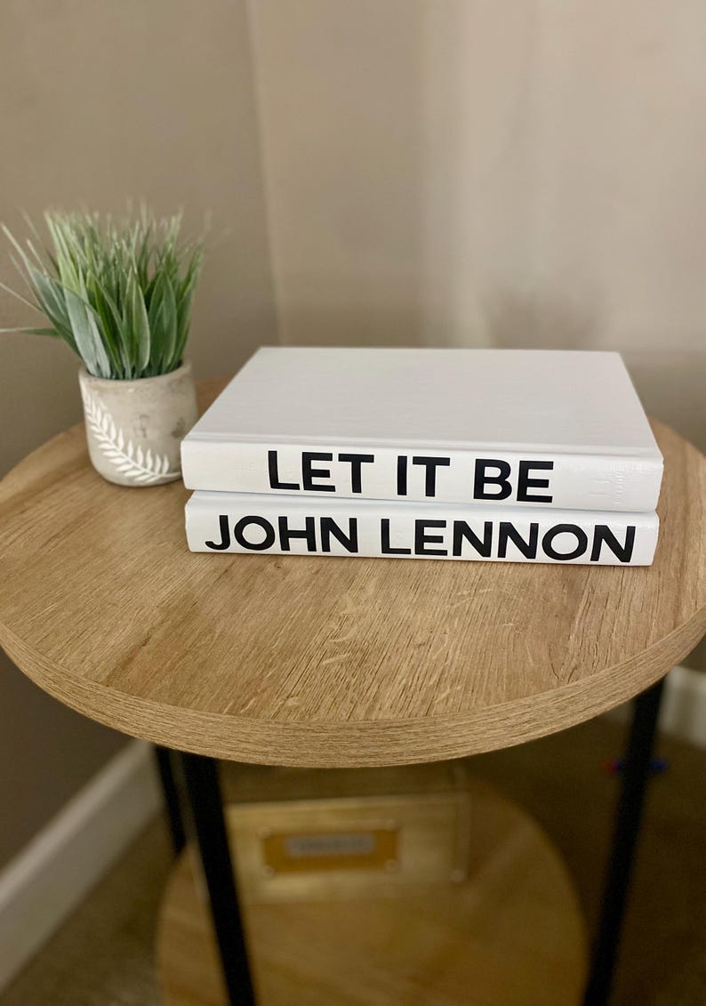 May include: Two white books stacked on a wooden table. The top book reads "LET IT BE" in black letters, and the bottom book reads "JOHN LENNON". A small potted plant with green grass sits to the left.