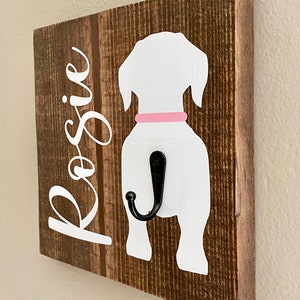 May include: Wooden dog leash holder with a white dog silhouette, a pink collar, and a black hook. The name "Rosie" is written in white script on the left side of the brown wooden board. The wood has a rustic, textured appearance.
