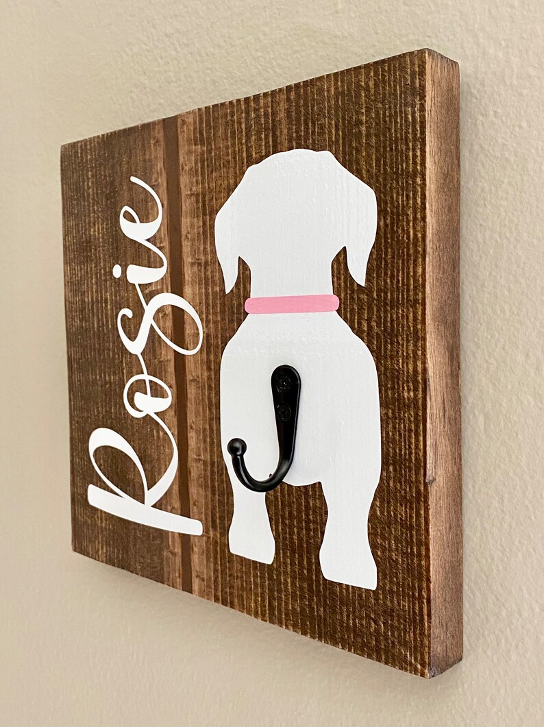 May include: Wooden dog leash holder with a white dog silhouette, a pink collar, and a black hook. The name "Rosie" is written in white script on the left side of the brown wooden board. The wood has a rustic, textured appearance.