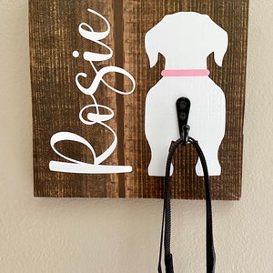 May include: A wooden dog leash holder with a white dog silhouette and the name "Rosie" in white script. The dog has a pink collar and a black hook for hanging leashes. A black leash hangs from the hook.