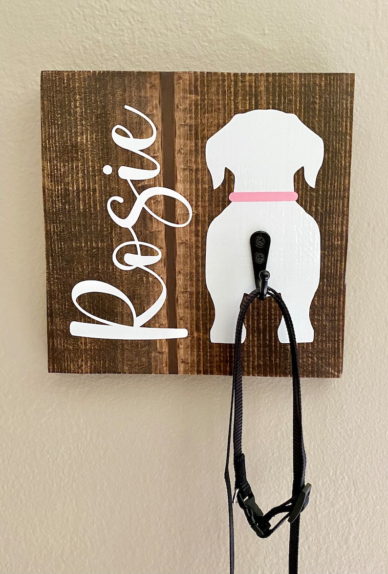 May include: A wooden dog leash holder with a white dog silhouette and the name "Rosie" in white script. The dog has a pink collar and a black hook for hanging leashes. A black leash hangs from the hook.