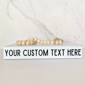 Peut inclure: Un livre blanc avec le texte "YOUR CUSTOM TEXT HERE" imprimé en noir sur le dos. Un brin de perles en bois repose sur le dessus du livre.