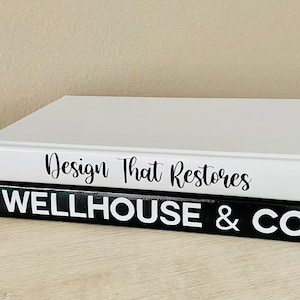May include: Two decorative books stacked on a light wood surface. The top book is white with the text "Design That Restores" in black script. The bottom book is black with the text "WELLHOUSE & CO" in white block letters.