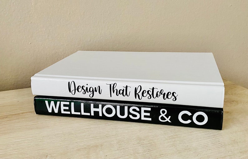 May include: Two decorative books stacked on a light wood surface. The top book is white with the text "Design That Restores" in black script. The bottom book is black with the text "WELLHOUSE & CO" in white block letters.