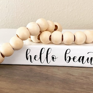 May include: A white book with the words "hello beautiful" in black script is adorned with a strand of natural wooden beads. The beads are light brown and round, creating a decorative accent.