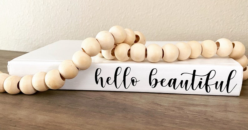 May include: A white book with the words "hello beautiful" in black script is adorned with a strand of natural wooden beads. The beads are light brown and round, creating a decorative accent.