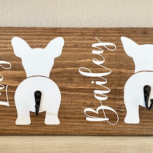 May include: Wooden wall-mounted key holder with a brown finish. Features two white dog silhouettes with black hooks. The names "Lucy" and "Bailey" are written in cursive script on the wood.