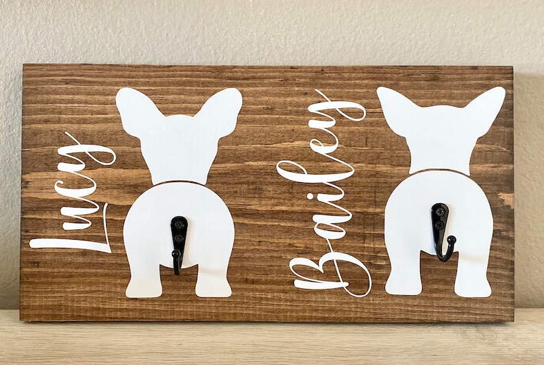 May include: Wooden wall-mounted key holder with a brown finish. Features two white dog silhouettes with black hooks. The names "Lucy" and "Bailey" are written in cursive script on the wood.