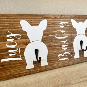 May include: A wooden wall-mounted dog leash holder with a rustic brown finish. Features two white dog silhouettes with black hooks and the names "Lucy" and "Bailey" in white script. A charming and functional home decor item.