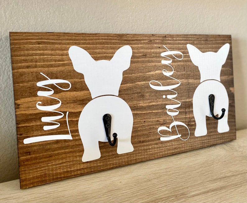 May include: A wooden wall-mounted dog leash holder with a rustic brown finish. Features two white dog silhouettes with black hooks and the names "Lucy" and "Bailey" in white script. A charming and functional home decor item.