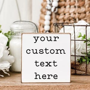 Puede incluir: Un letrero de bloque de madera blanco con el texto "your custom text here" en letras negras. El letrero tiene un borde marrón y se muestra sobre una superficie de madera. Los elementos decorativos incluyen flores blancas, un tarro de cristal y una cesta de alambre.