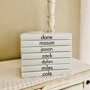 May include: A stack of white books with black text displaying names: Dane, Mason, Jaxon, Zack, Dylan, Miles, and Cole. The books are stacked on a white surface with a textured pattern, and a decorative white candlestick is in the background.