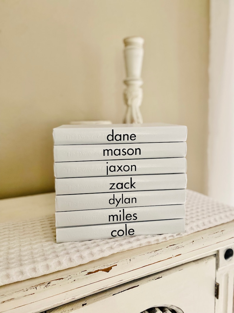 May include: A stack of white books with black text displaying names: Dane, Mason, Jaxon, Zack, Dylan, Miles, and Cole. The books are stacked on a white surface with a textured pattern, and a decorative white candlestick is in the background.
