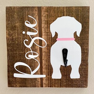 May include: A wooden dog leash holder with a white dog silhouette and a black hook. The dog has a pink collar. The name "Rosie" is written in white cursive on the left side of the brown wooden board.