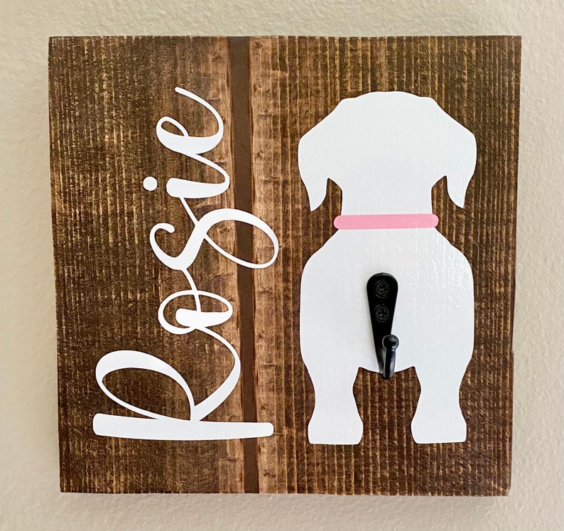 May include: A wooden dog leash holder with a white dog silhouette and a black hook. The dog has a pink collar. The name "Rosie" is written in white cursive on the left side of the brown wooden board.