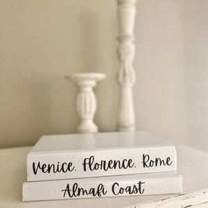 May include: Two white decorative books with black text that reads "Venice, Florence, Rome" and "Amalfi Coast." Two white decorative candle holders are behind the books. The books and candle holders are on a white surface.