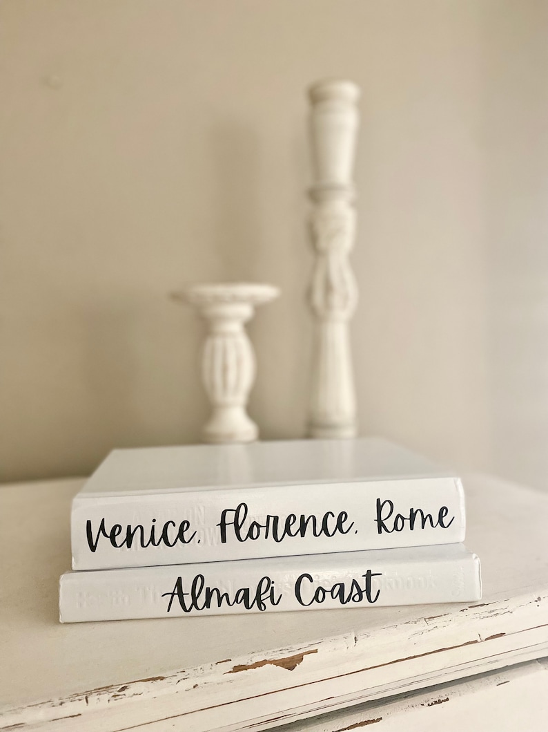 May include: Two white decorative books with black text that reads "Venice, Florence, Rome" and "Amalfi Coast." Two white decorative candle holders are behind the books. The books and candle holders are on a white surface.