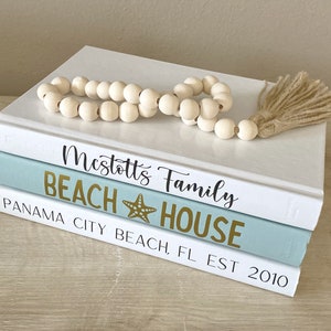 May include: Three decorative books stacked on a light-colored surface. The top book is white, the middle book is light blue, and the bottom book is white. The middle book has the words "BEACH HOUSE" and a starfish graphic in gold. A wooden bead garland with a tassel rests on top.