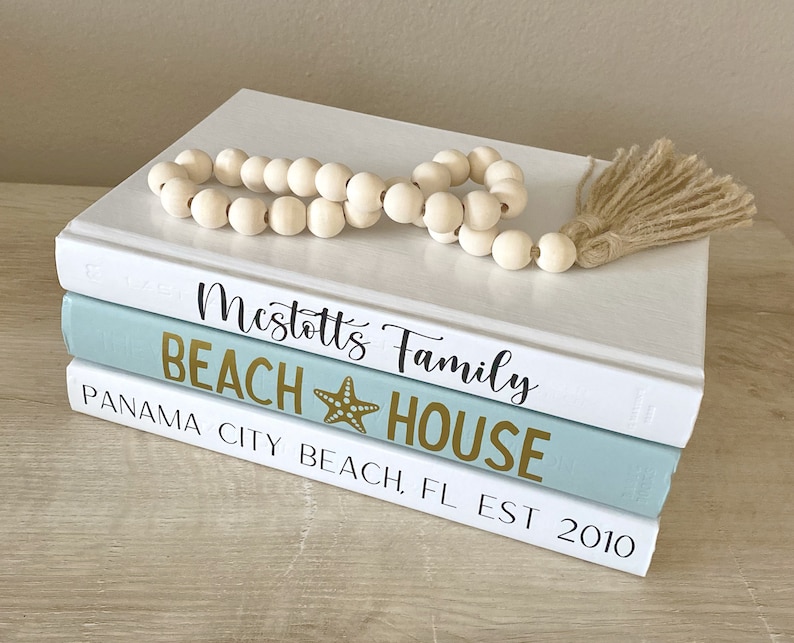 May include: Three decorative books stacked on a light-colored surface. The top book is white, the middle book is light blue, and the bottom book is white. The middle book has the words "BEACH HOUSE" and a starfish graphic in gold. A wooden bead garland with a tassel rests on top.