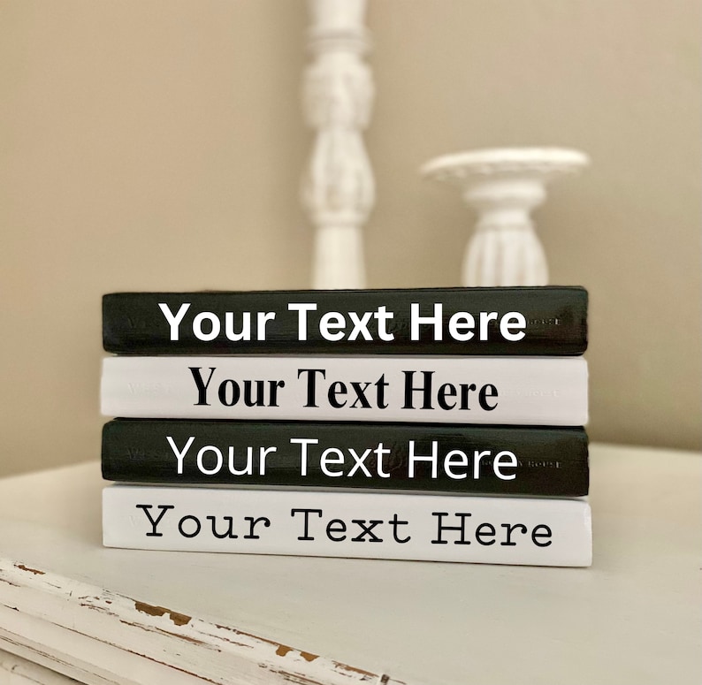 May include: A stack of four decorative books in black and white. Each book has the text "Your Text Here" printed in a bold, sans-serif font. The books are arranged on a white surface, with a blurred background.