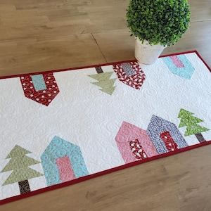 May include: A rectangular, quilted table runner with a white background and a red border. The design features colorful houses and trees in shades of blue, pink, red, and green. The runner is placed on a wooden table.