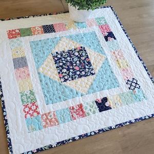 May include: A square quilted table runner with a floral and geometric pattern. The center features a light blue diamond shape with a dark blue floral square. The border has colorful squares and a white background with a navy floral edge.