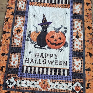 May include: A Halloween-themed quilt with an orange and black color scheme. The quilt features a black cat wearing a witch's hat, pumpkins, and the text "Happy Halloween".