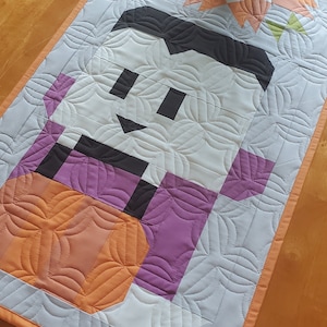 May include: A quilted table runner with a whimsical design featuring a white ghost holding a purple and orange pumpkin. The runner has a light gray background with a repeating pattern of curved lines.
