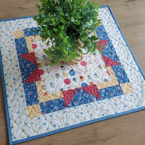 May include: A blue and white quilted table runner with a red, yellow, and blue floral pattern in the center. The runner has a blue border and is decorated with a quilted leaf design.