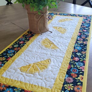 May include: A rectangular quilted table runner featuring a central white panel with yellow lemon slice appliques and stippled stitching. The border is a dark navy fabric with a colorful floral pattern, finished with a bright yellow fabric binding.