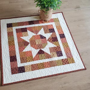 May include: A square quilted table topper featuring a central star motif surrounded by a border of small patchwork squares in shades of orange, brown, and gold. The quilt has white fabric sections with intricate swirling machine quilting patterns.
