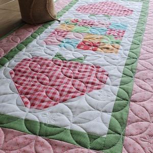 May include: A quilted table runner featuring a strawberry design. The runner has a pink background with a green border and white accents. The strawberries are red and white gingham. The runner is approximately 40 inches long.